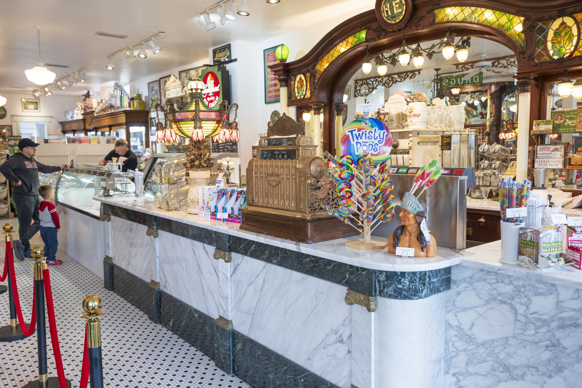 A vintage-style soda fountain and candy shop with a marble countertop, brass cash register, and colorful lollipops on display. The shop features ornate woodwork, antique décor, and classic stained glass accents. A staff member serves ice cream behind the counter while a customer and a young child wait. The nostalgic atmosphere is reminiscent of early 20th-century soda fountains.