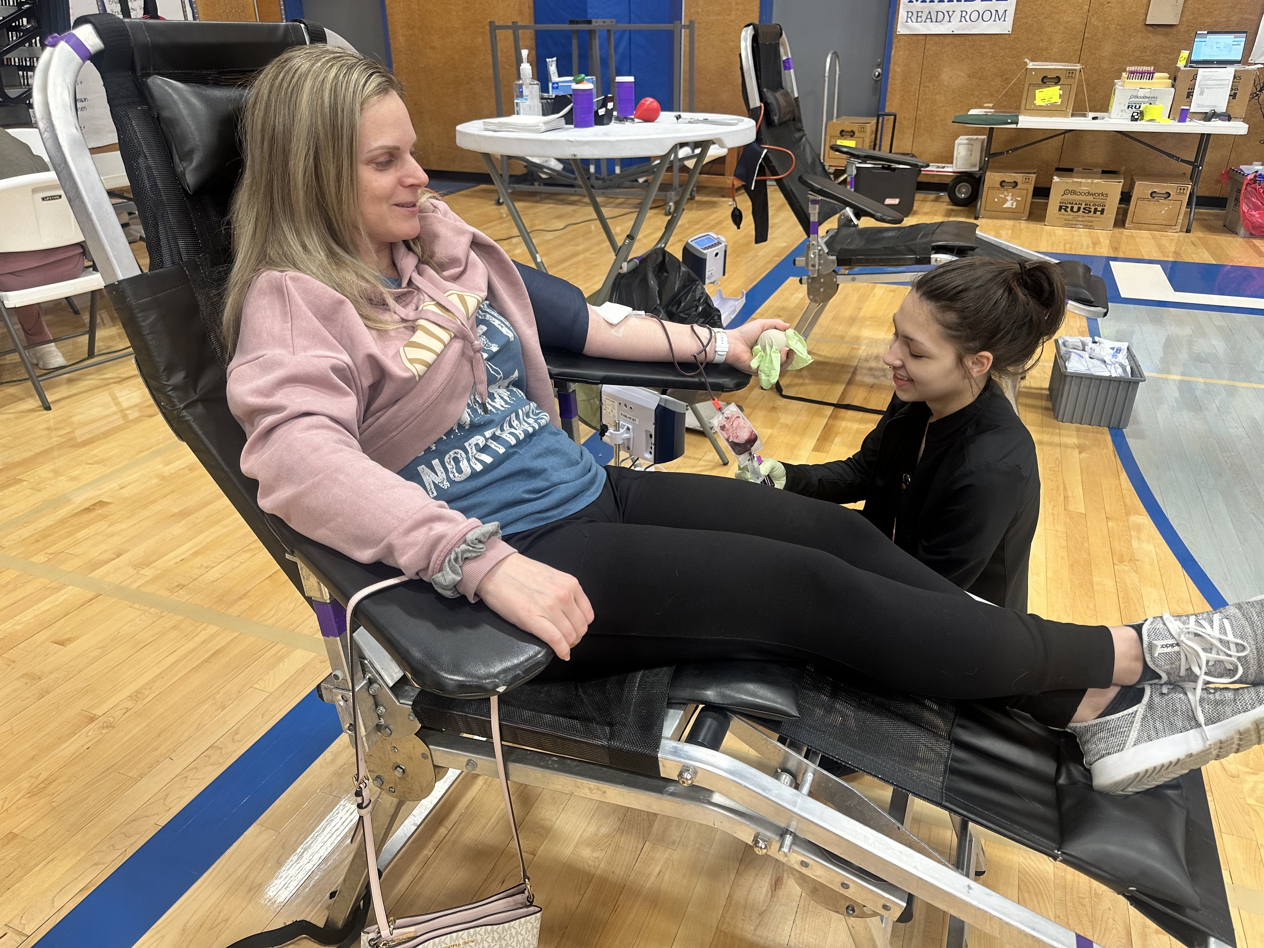 Inside a school gym, a woman is lying on a reclined chair while a healthcare professional in gloves assists with the donation process.